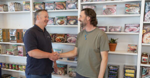 Two people shake hands in front of shelves stocked with packaged food items, including boxed meals, canned goods, and jars, suggesting a friendly or professional interaction in a store or product display area.
