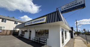 A single?story white?brick laundromat with large front windows showing washers and dryers inside. Two signs display the name “The Fold Laundromat,” one on the building and one elevated above it with the tagline “All Welcome, Always.” Two black benches and a hanging flower basket sit near the entrance under a mostly clear sky.