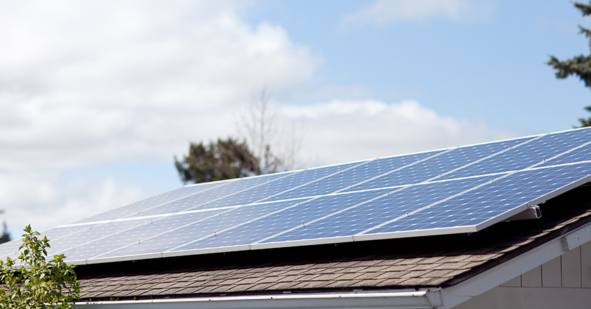 “A set of rectangular solar panels installed on a brown?shingled roof, arranged in angled rows beneath a cloudy sky. A tree canopy is visible behind the house.”