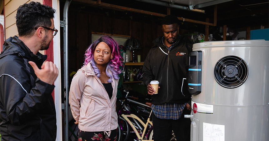 Three people stand in a garage beside a large cylindrical water heater or heat?pump unit. One person gestures while speaking, another listens closely, and a third holds a cup while observing. Bicycles and typical garage storage items are visible in the background.