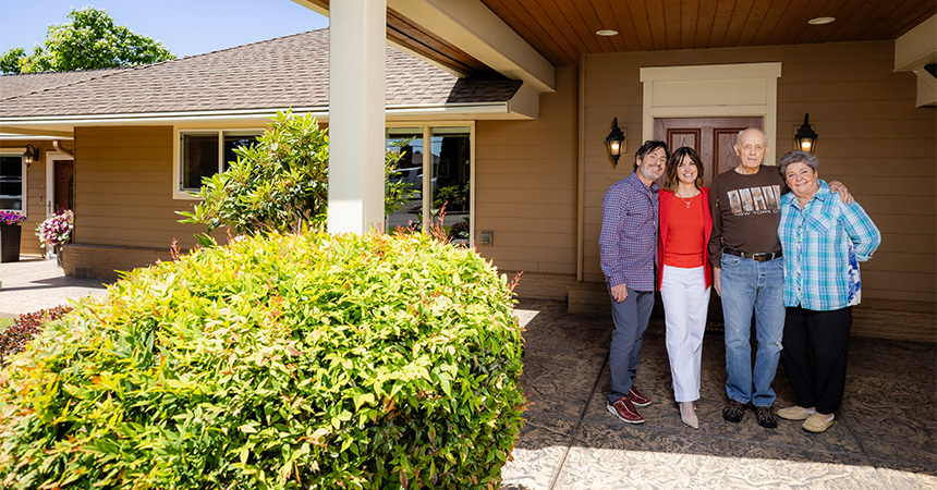 Four people standing together on a covered porch in front of a tan house with white trim and a wooden front door, with shrubs and potted flowers nearby.