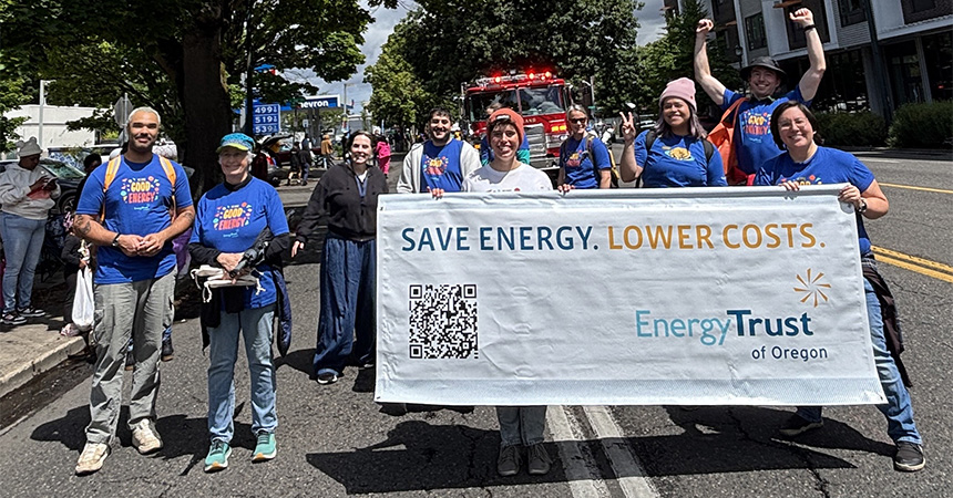 A group of people in blue shirts stand on a sunny street holding a large banner that reads “SAVE ENERGY. LOWER COSTS. Energy Trust of Oregon,” with a QR code printed on it. A fire truck and other parade participants are visible behind them, along with trees and buildings in the background.