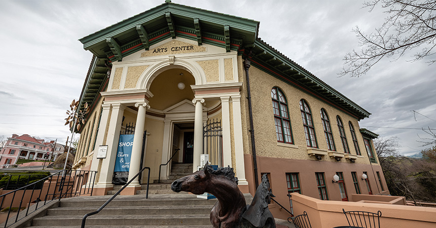A historic arts center building with tall columns, arched windows, and a green roof. A staircase leads to the entrance, where a sign advertises arts, crafts, and gifts. A sculpted horse head stands near the bottom of the steps.