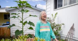 Resident standing beside a tall sunflower in a garden next to a white manufactured home and backyard shed.