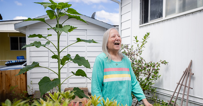Resident standing beside a tall sunflower in a garden next to a white manufactured home and backyard shed.