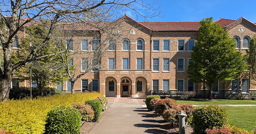 A three-story brick institutional building stands at the end of a landscaped walkway on a clear, sunny day. The symmetrical building features a red tile roof, rows of large windows, and an arched main entrance centered on the façade. Shrubs, flowering plants, and mature trees line the path leading to the entrance, with neatly maintained lawns on either side. The image highlights the building's historic architectural style and well-kept campus setting under a bright blue sky.