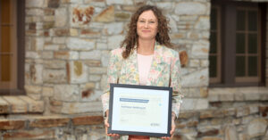 A person stands outdoors in front of a stone building, holding a framed certificate with both hands. The person is wearing a light-colored floral-patterned blazer over a pale shirt. The certificate, displayed in a black frame, includes the year “2026” and appears to be an award or recognition document. Two windows and a textured stone wall form the background. The image is warmly lit and focused on the award recipient and the framed certificate.