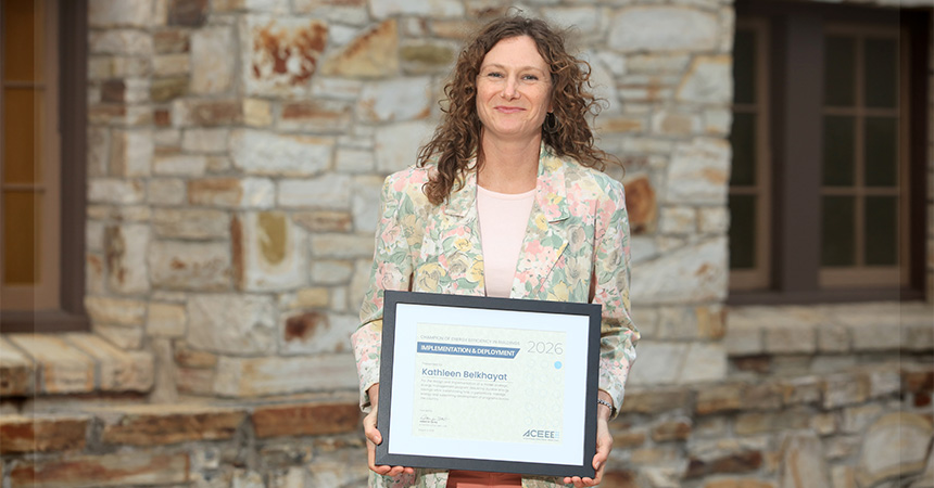 A person stands outdoors in front of a stone building, holding a framed certificate with both hands. The person is wearing a light-colored floral-patterned blazer over a pale shirt. The certificate, displayed in a black frame, includes the year “2026” and appears to be an award or recognition document. Two windows and a textured stone wall form the background. The image is warmly lit and focused on the award recipient and the framed certificate.