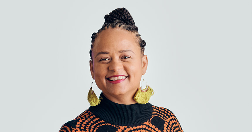 Studio portrait of a person with braided hair against a light gray background. The individual is wearing a black turtleneck sweater with an orange geometric pattern and large tassel earrings, facing the camera in a head-and-shoulders composition.