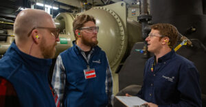 Three workers wearing safety glasses and earplugs stand inside an industrial mechanical room, discussing equipment. Large pipes, valves, and a cylindrical machine fill the background while one worker holds a clipboard during the conversation.