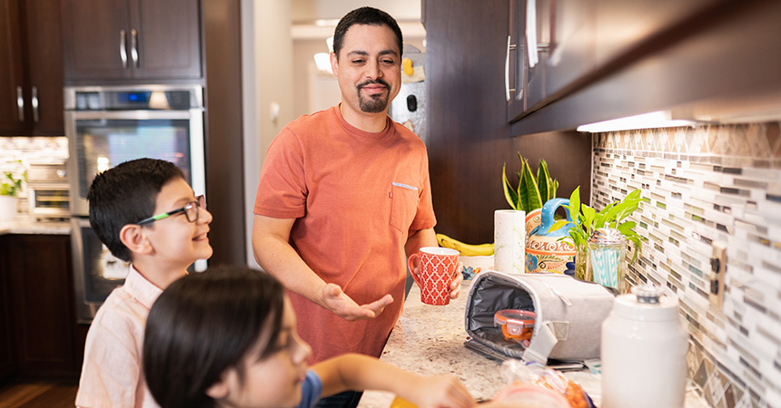 A person stands in a modern kitchen beside two children near a countertop with a lunch bag, food containers, and a reusable water bottle. Dark wood cabinets, a tiled backsplash, and built-in appliances are visible in the background, creating a warm, home kitchen setting.