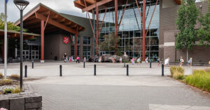 The exterior of the Salvation Army Kroc Center, featuring a large modern building with timber architectural elements and expansive windows. A group of children and adults walk across the plaza in front of the entrance.