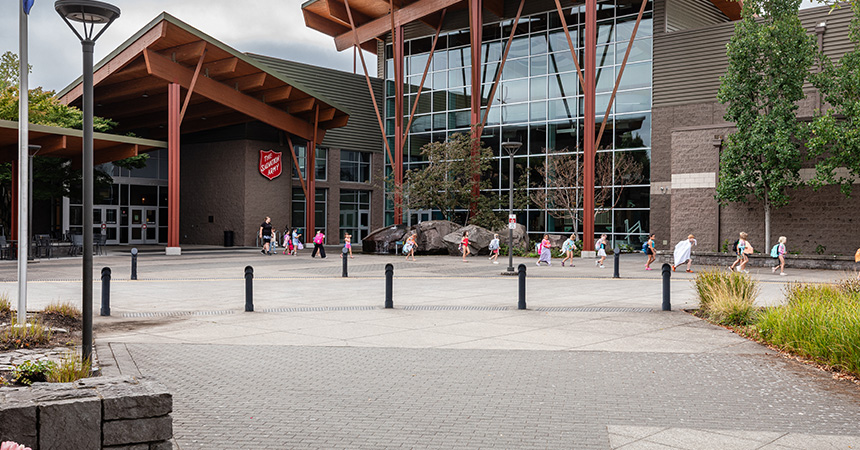 The exterior of the Salvation Army Kroc Center, featuring a large modern building with timber architectural elements and expansive windows. A group of children and adults walk across the plaza in front of the entrance.