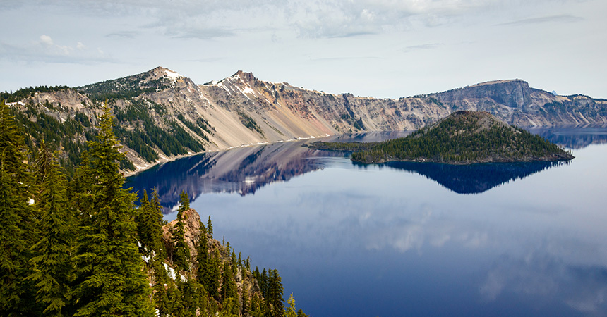 Wide view of a deep blue volcanic crater lake surrounded by steep rocky cliffs and evergreen forests. A small forested island sits near the center of the calm water, which reflects the surrounding mountains and sky. Trees frame the foreground, and light clouds hang above the rugged landscape.